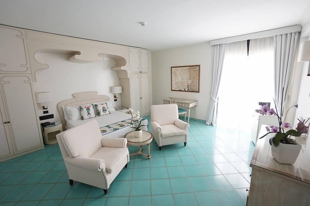 A hotel room with a bed, two armchairs, a small table, a desk, and a potted plant.