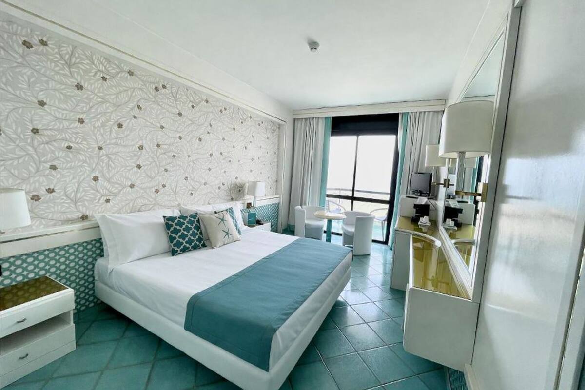 A hotel room with a large bed, a desk, a chair, a small table, and a bathroom area.