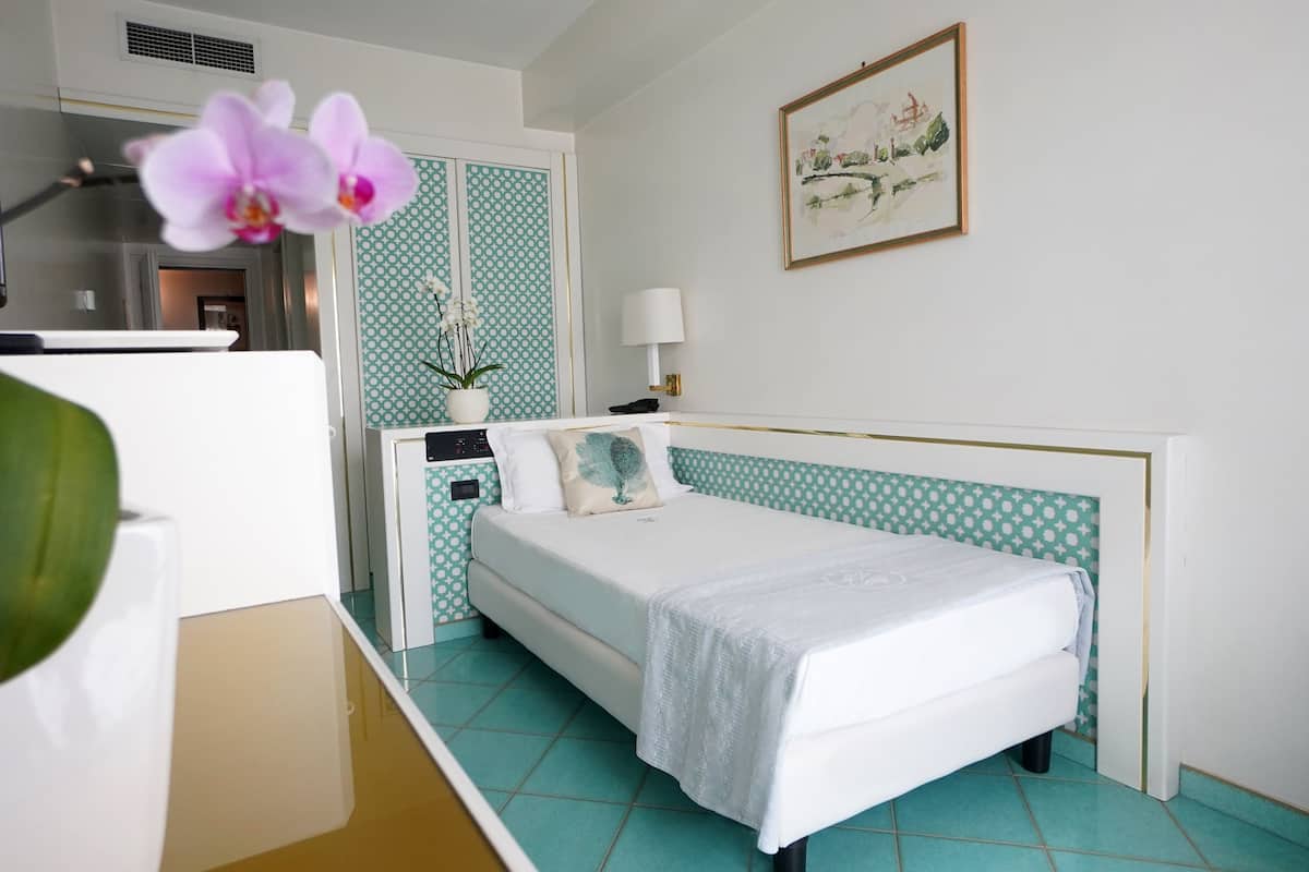 A neatly arranged hotel room with a bed, a desk, a potted orchid, and a framed artwork on the wall.