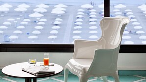 A white chair and table with a glass of orange juice and a newspaper by a window overlooking a beach with umbrellas. - Grand Hotel Terme (Margherita di Savoia)
