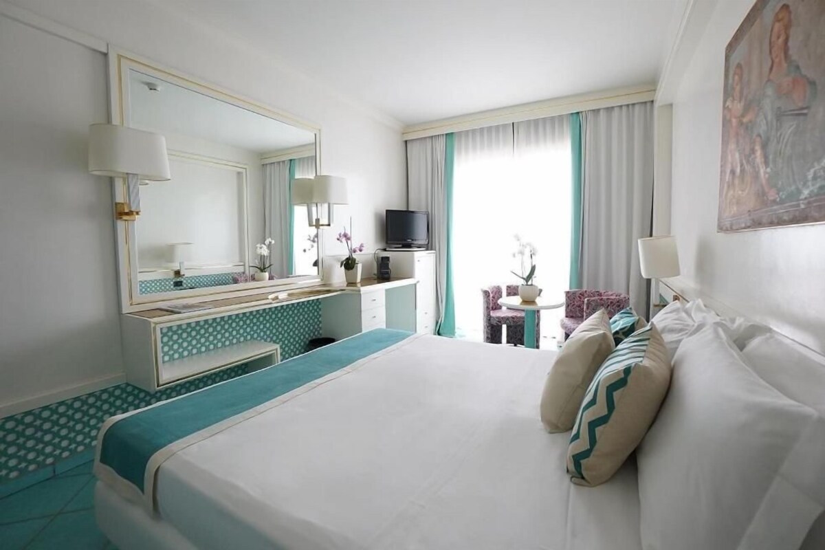 A hotel room with a large bed, a desk with a TV, a mirror, and a window with curtains.