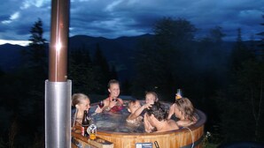 A group of people in a wooden hot tub with a metal pole, surrounded by trees and mountains in the background. - Blue Mountain Hotel (Treffen)