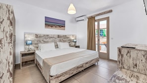 A bedroom with a bed, bedside lamps, a wall-mounted air conditioner, a painting, and a view through a door. - Erato Hotel (Hersonissos)