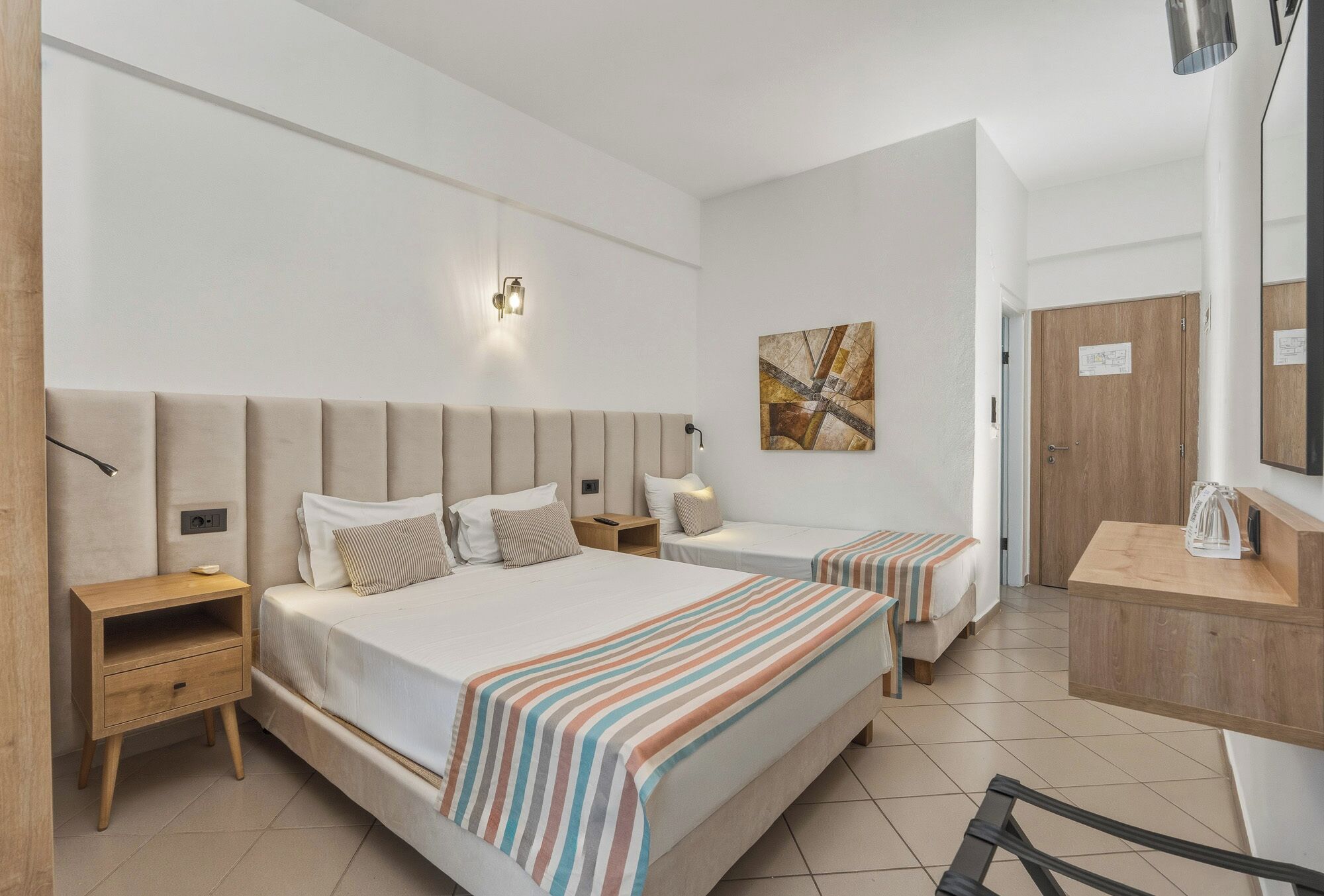 A hotel room with two beds, a bedside table, a nightstand, a wooden dresser, a wall-mounted lamp, and a painting.