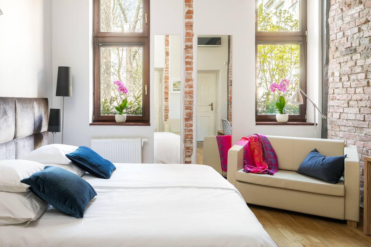 A bedroom with a bed, a sofa, and two windows with plants.