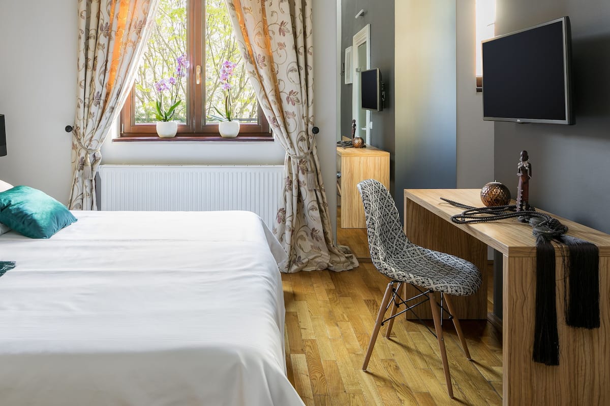A bedroom with a bed, a desk, a chair, a window with curtains, and a TV on the wall.