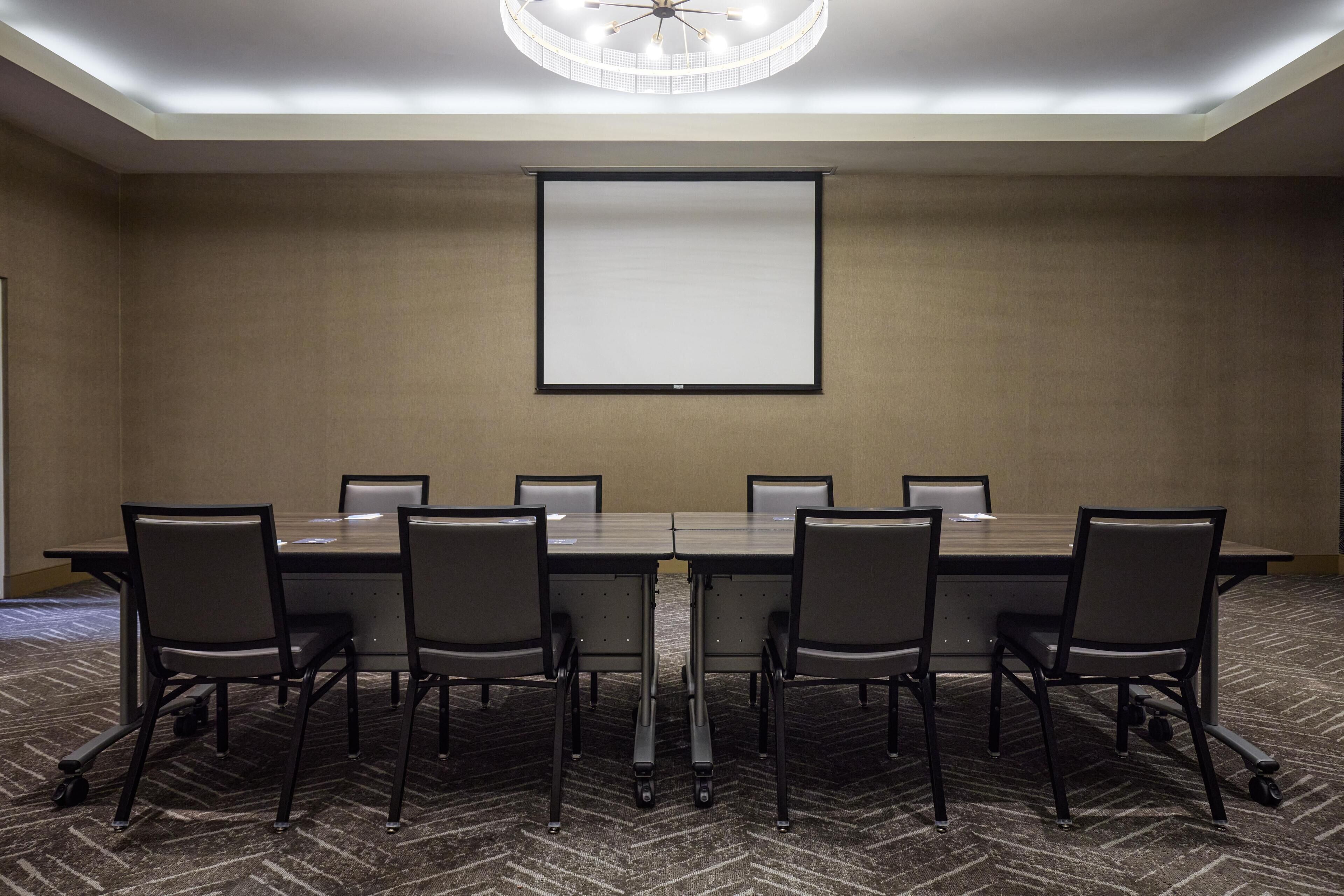 A conference room with a long table, six chairs, a projector screen, and a carpeted floor.