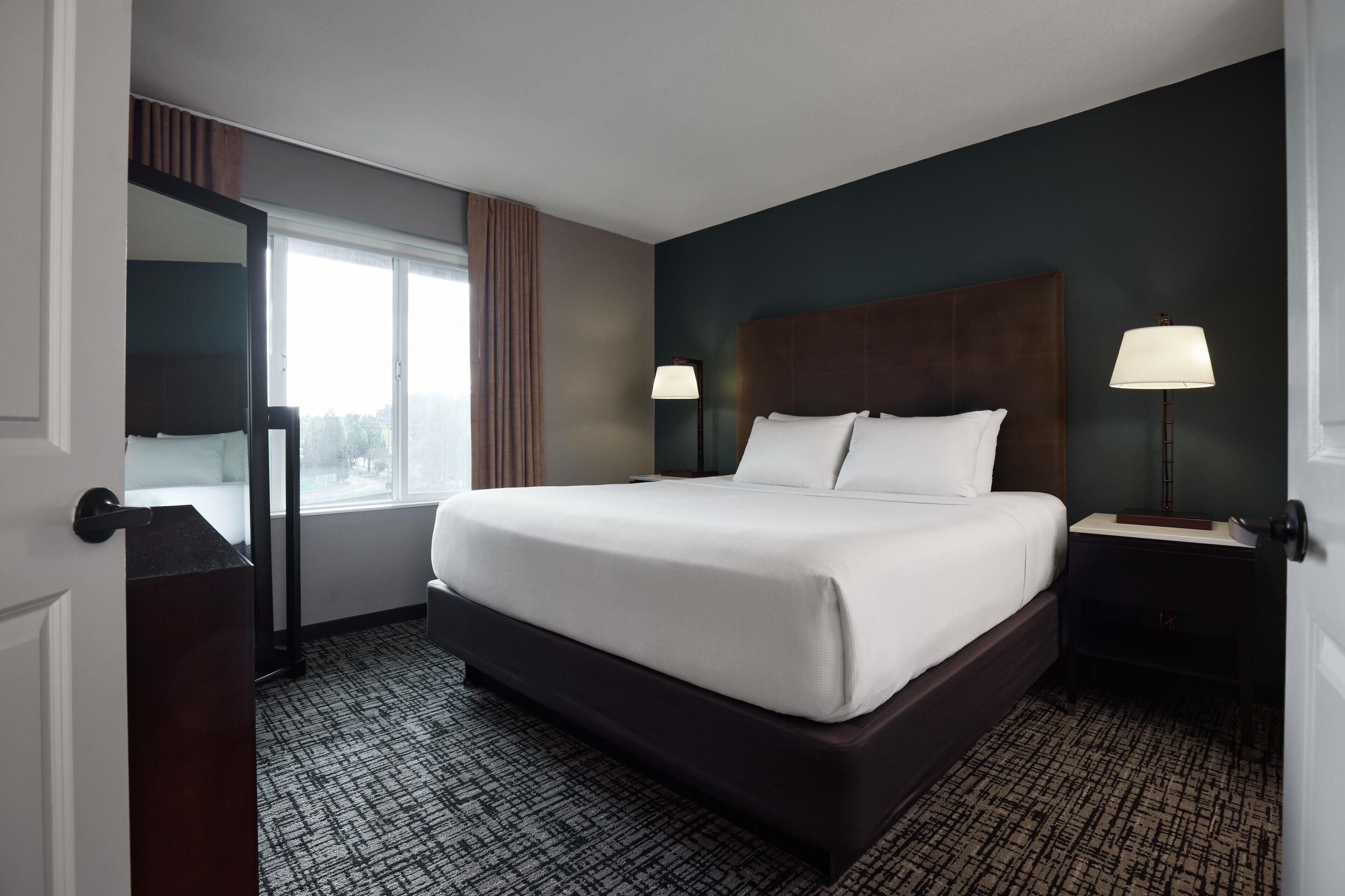 A hotel room with a large bed, two bedside tables with lamps, a dark headboard, a large window with curtains, and a patterned carpet.