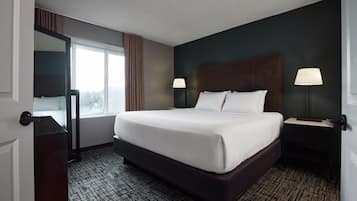 A hotel room with a large bed, two bedside tables with lamps, a dark headboard, a large window with curtains, and a patterned carpet.