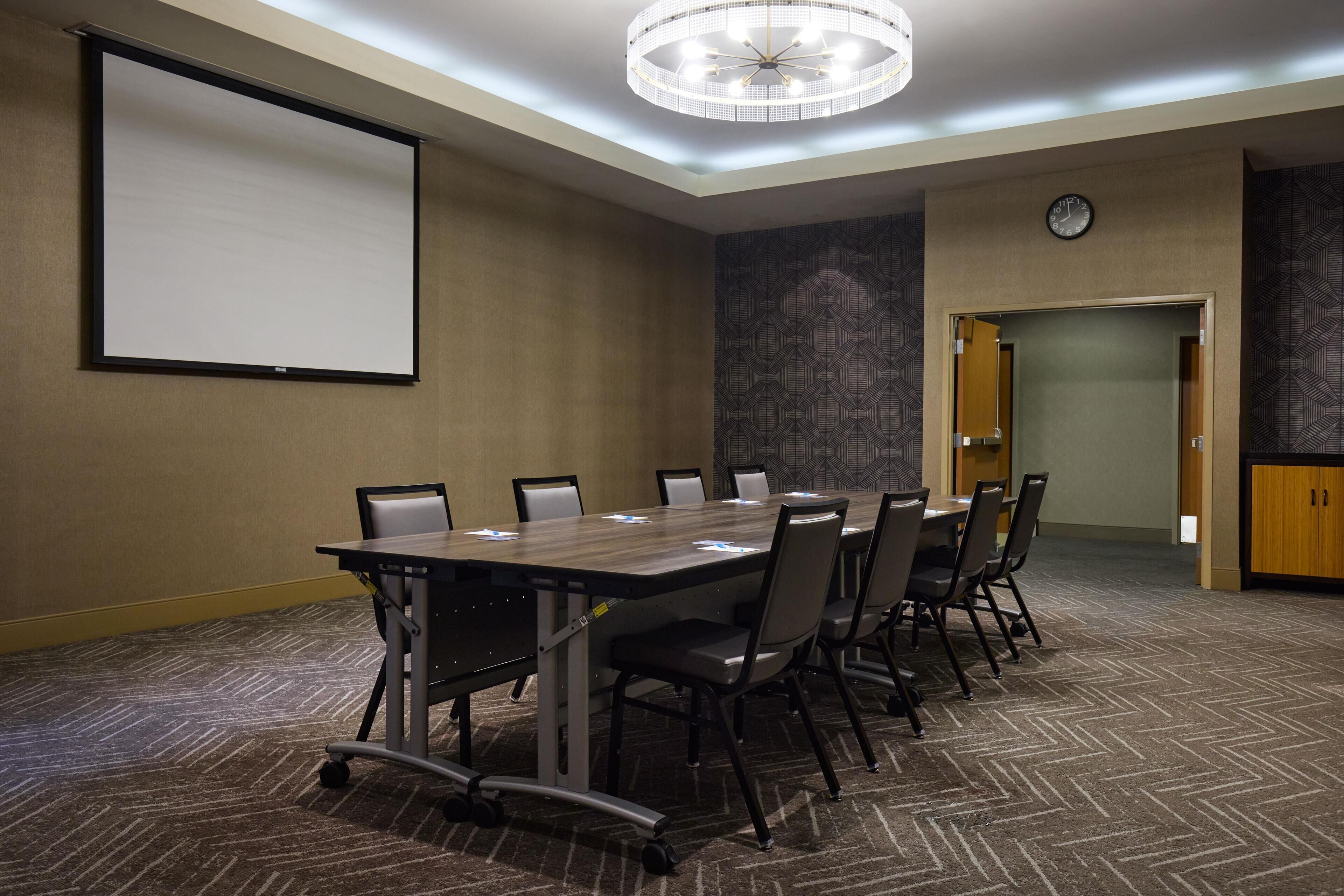 Une salle de conférence équipée d’une grande table, de chaises, d’un écran de projection, d’une horloge et d’un tapis à motifs.