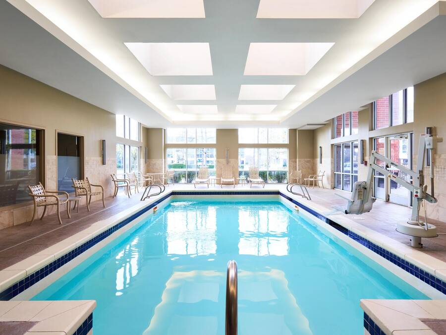 Una piscina interior con agua cristalina y azul, rodeada de sillones de descanso y un techo iluminado por el sol.