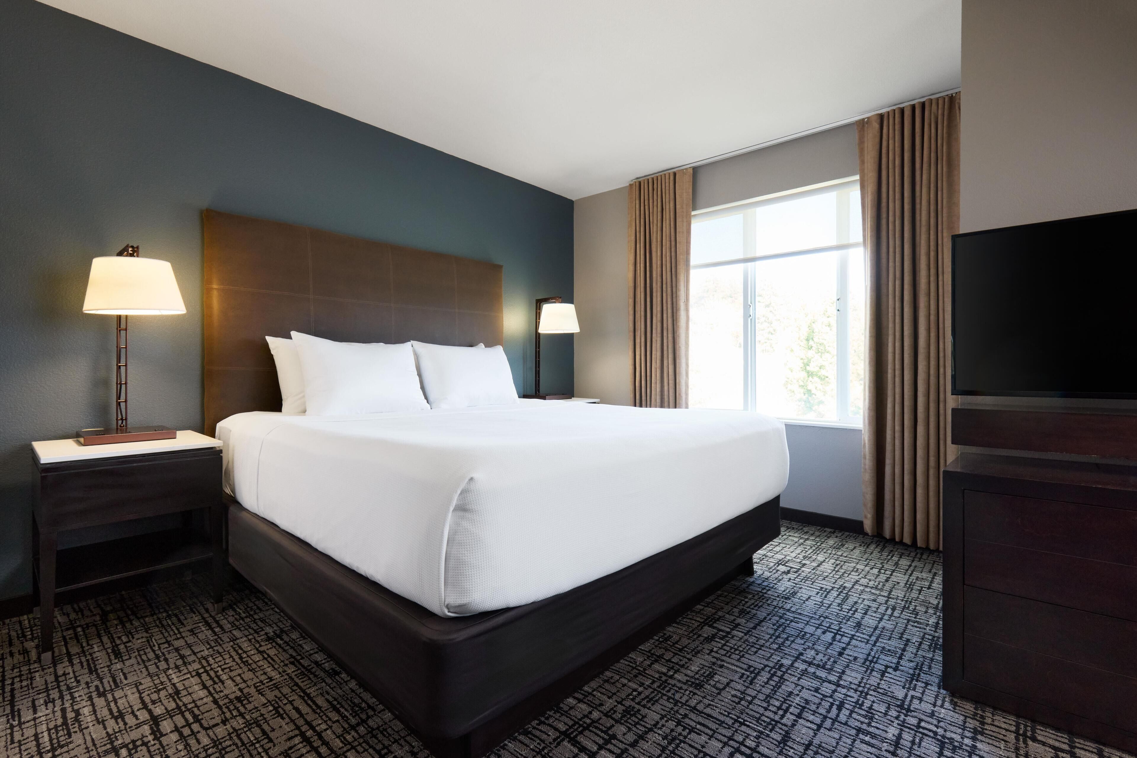 A hotel room with a large bed, two bedside tables with lamps, a flat-screen TV, and a window with curtains.