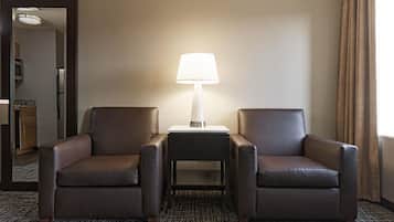 Habitación de hotel con dos sillones de cuero, una mesita con lámpara, alfombra estampada y vista a la cocina por un vano abierto.