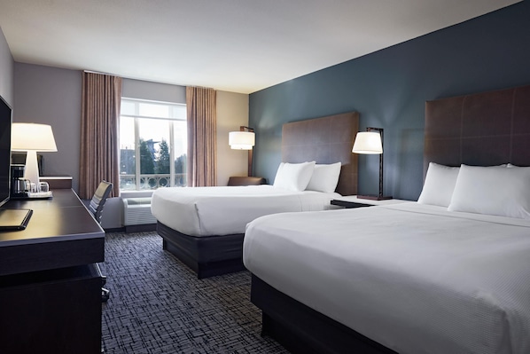 Quarto de hotel com duas camas, uma escrivaninha, uma luminária e uma janela com cortinas.