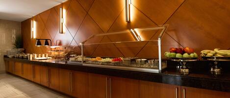 Uma área de buffet com parede em painel de madeira, vitrine de vidro para alimentos e uma variedade de frutas e legumes.