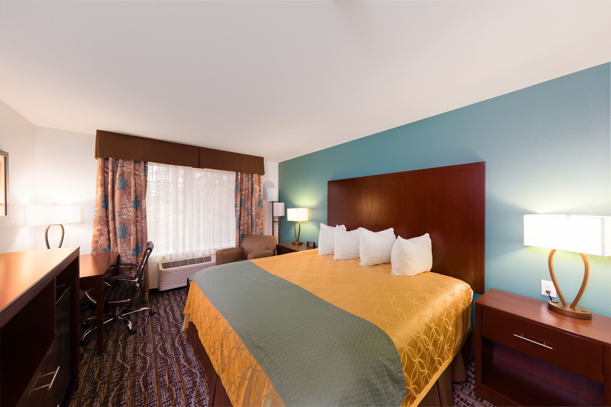 A hotel room with a large bed, a desk, a chair, a nightstand, and a lamp.
