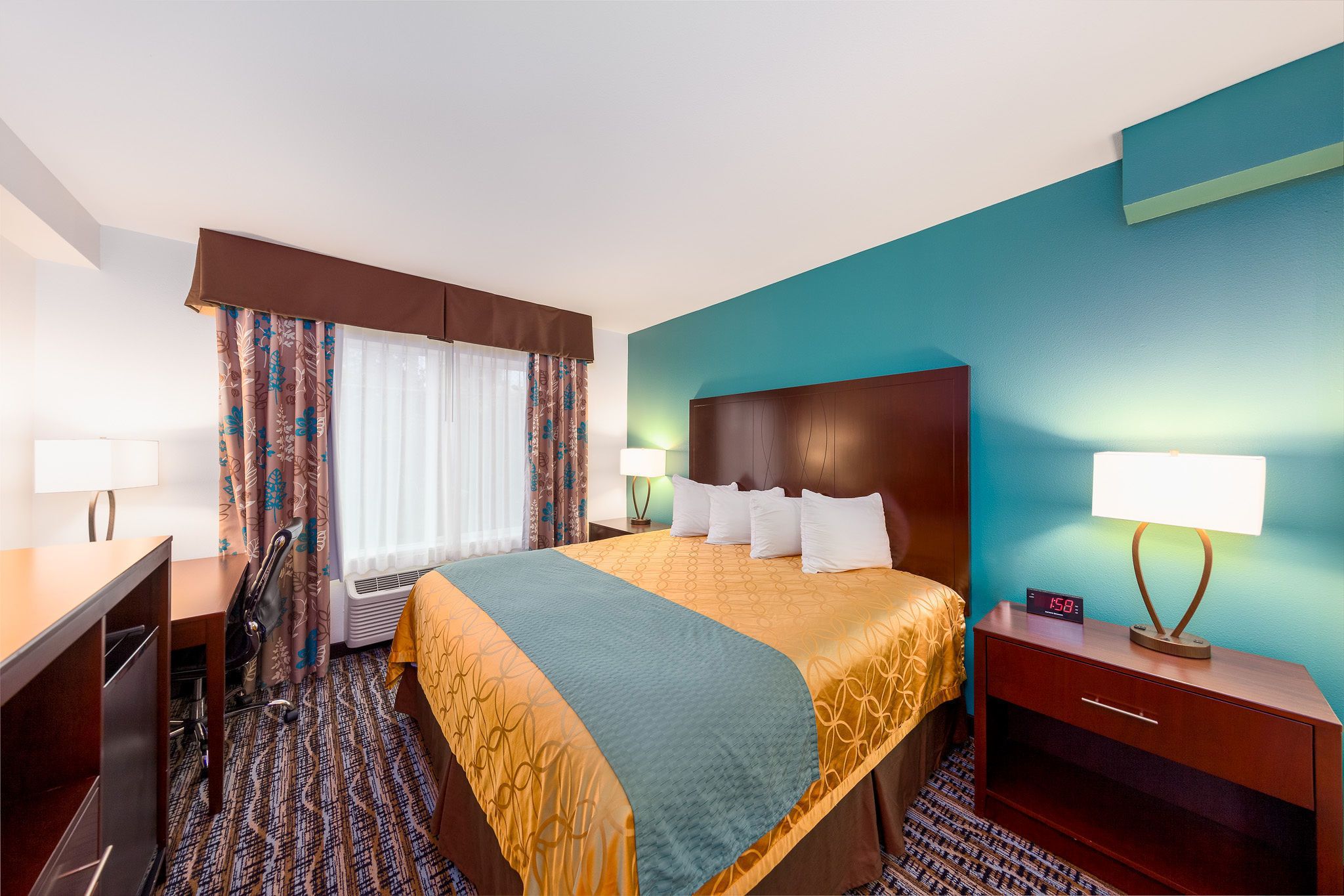 A hotel room with a bed, a desk, a chair, a lamp, and a clock.