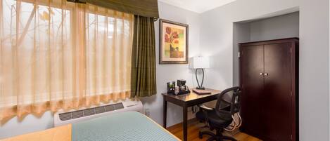A hotel room with a bed, desk, chair, wardrobe, and a painting on the wall.