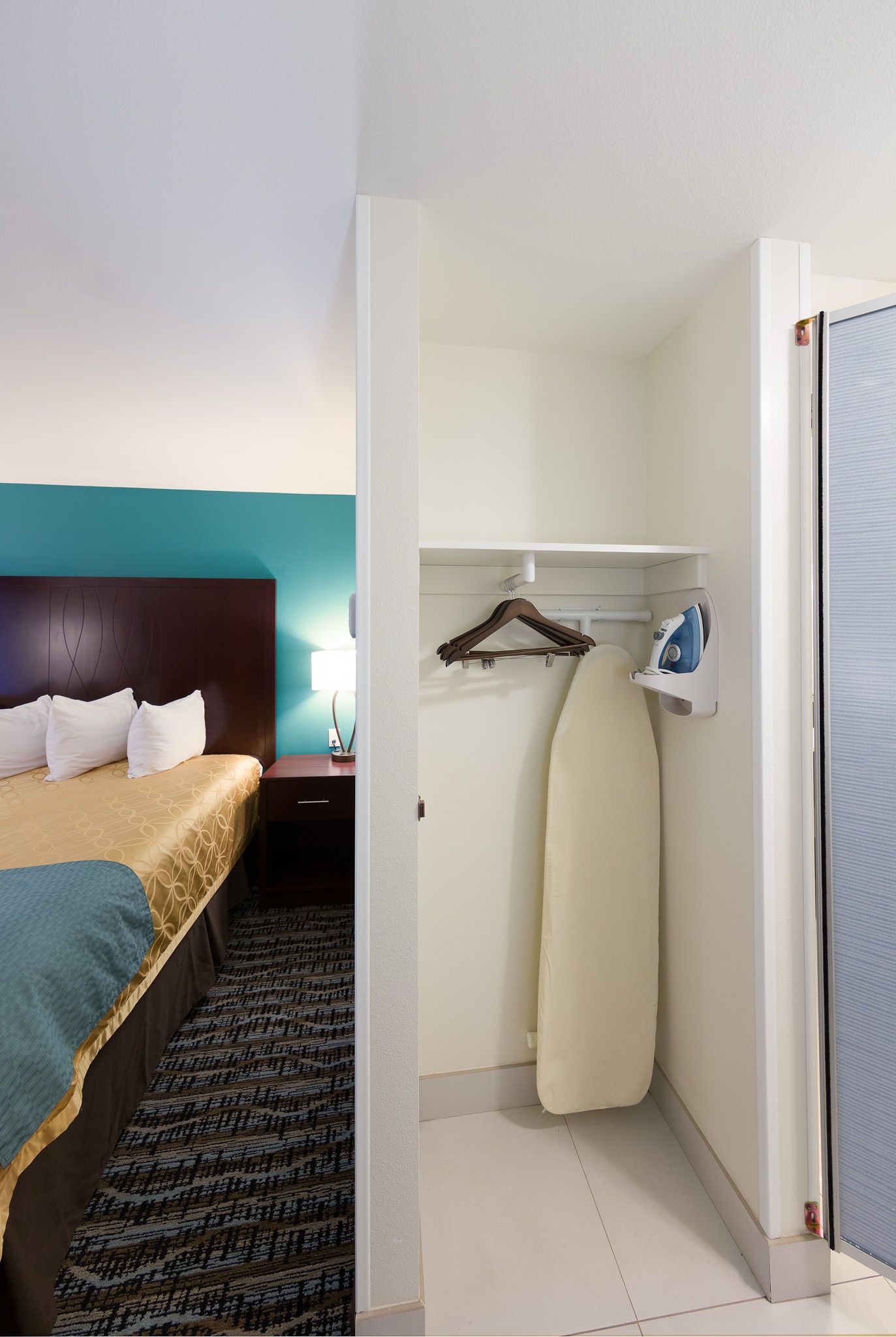 A hotel room with a bed, a nightstand, a wall-mounted ironing board, and a hanging clothes rack.
