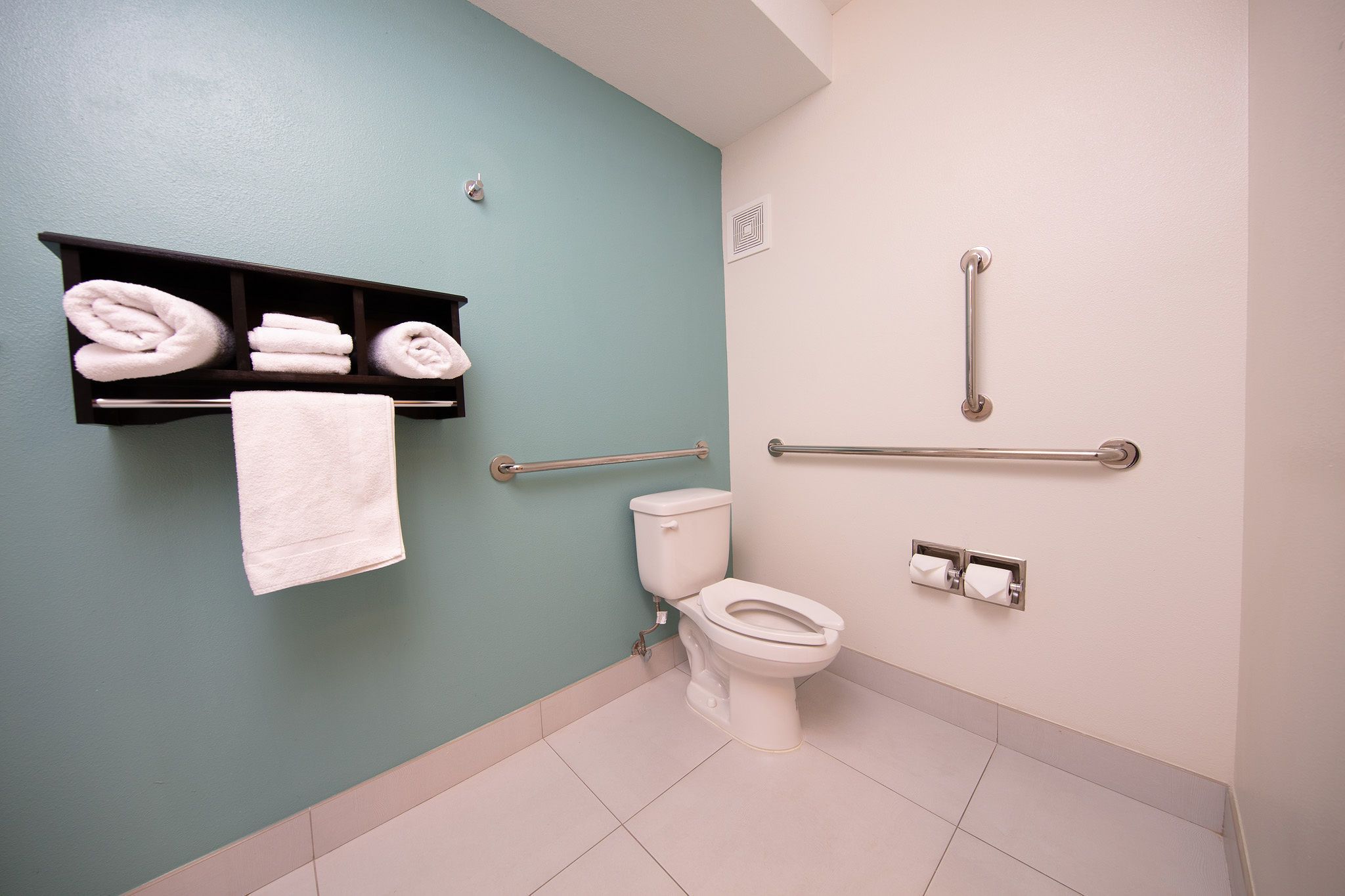 A bathroom with a teal wall, a white toilet, a towel rack with folded towels, and a grab bar.