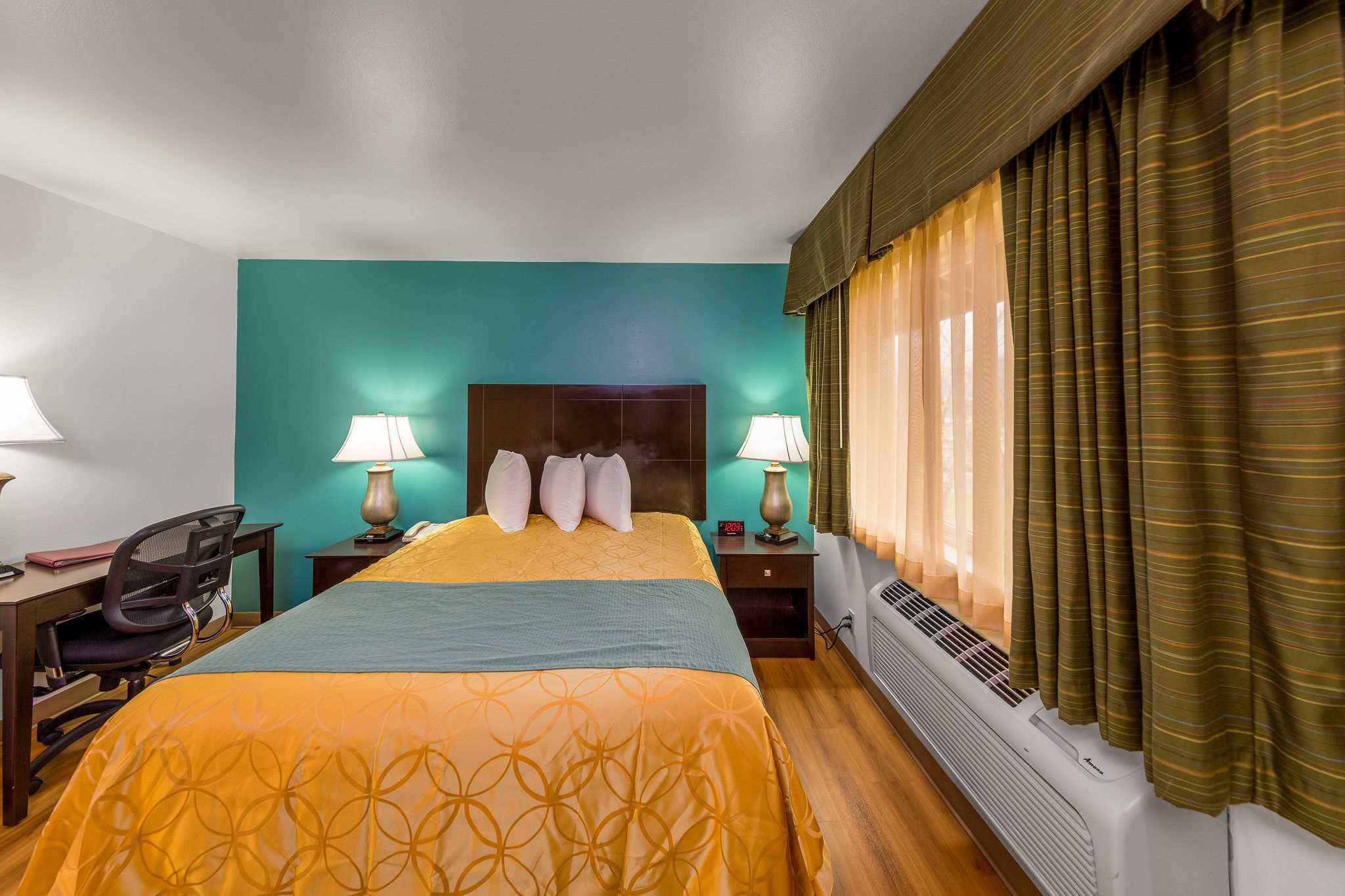 Une chambre d’hôtel comprenant un lit, deux lampes de chevet, un bureau avec une chaise, un radiateur et des rideaux.