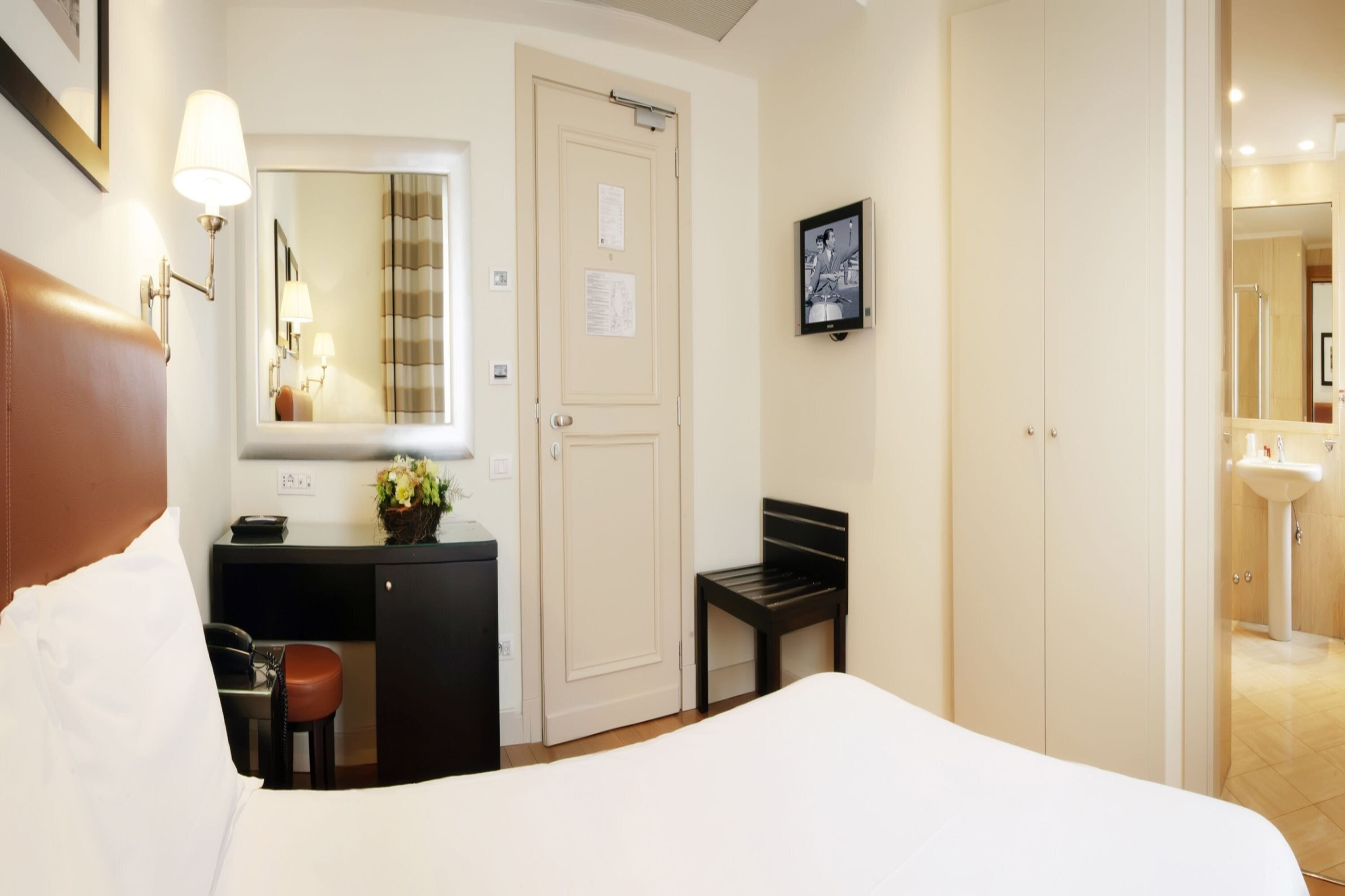 A hotel room with a bed, a desk with a telephone, a chair, a small table with a plant, a mirror, and a door.