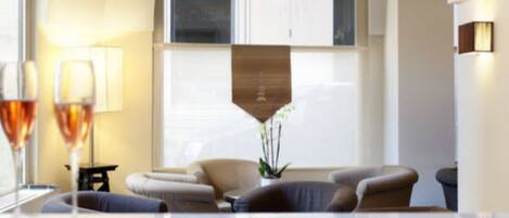 A modern hotel room with a large window, a sofa set, a small table with two glasses of wine, and a decorative plant.