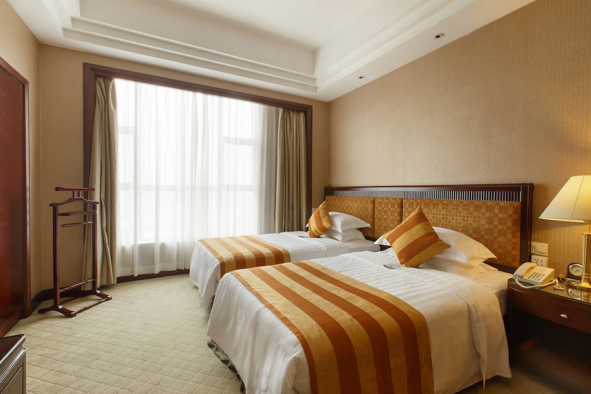 A hotel room with two beds, a bedside table with a lamp, a telephone, and a window with curtains.