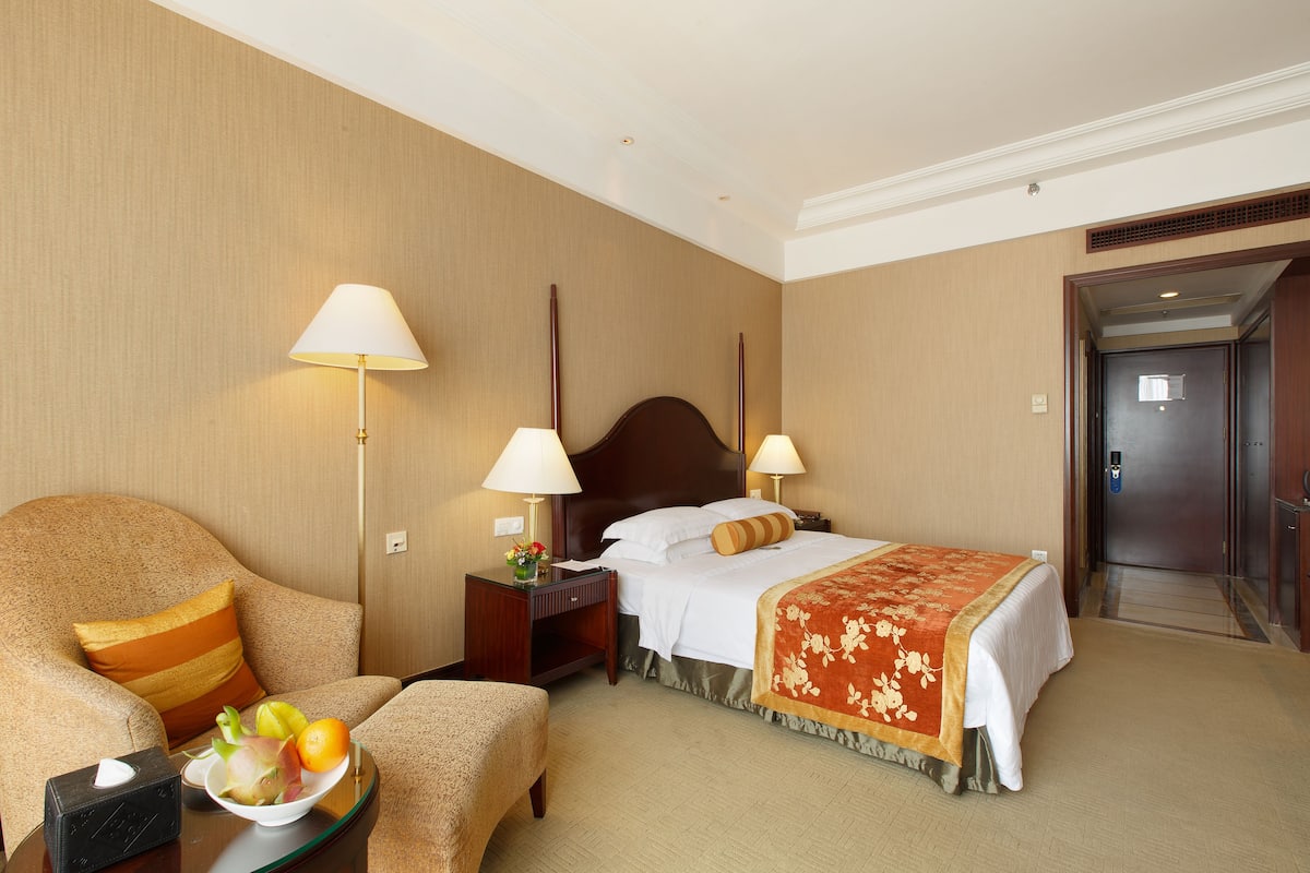 A hotel room with a bed, a sofa, a nightstand with a lamp, and a tray with fruits and a tissue box.