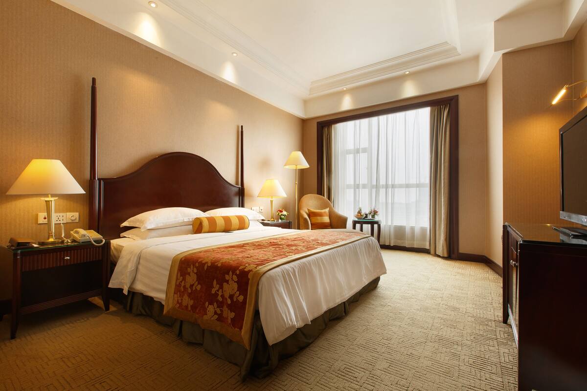 A hotel room with a large bed, two bedside tables with lamps, a chair, a television, and a large window with curtains.