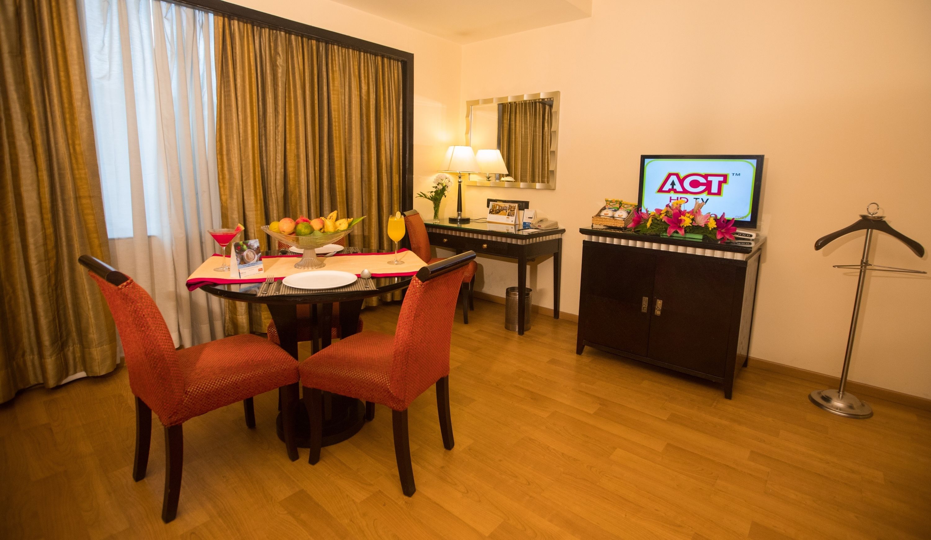 A hotel room with a dining table, two red chairs, a TV, a cabinet with flowers, and a floor lamp.