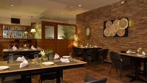 Un restaurant avec de nombreuses tables dressées pour le service, un bar bien garni de diverses bouteilles et un mur décoratif agrémenté d’un grand miroir rond et d’une œuvre d’art à la texture intéressante.