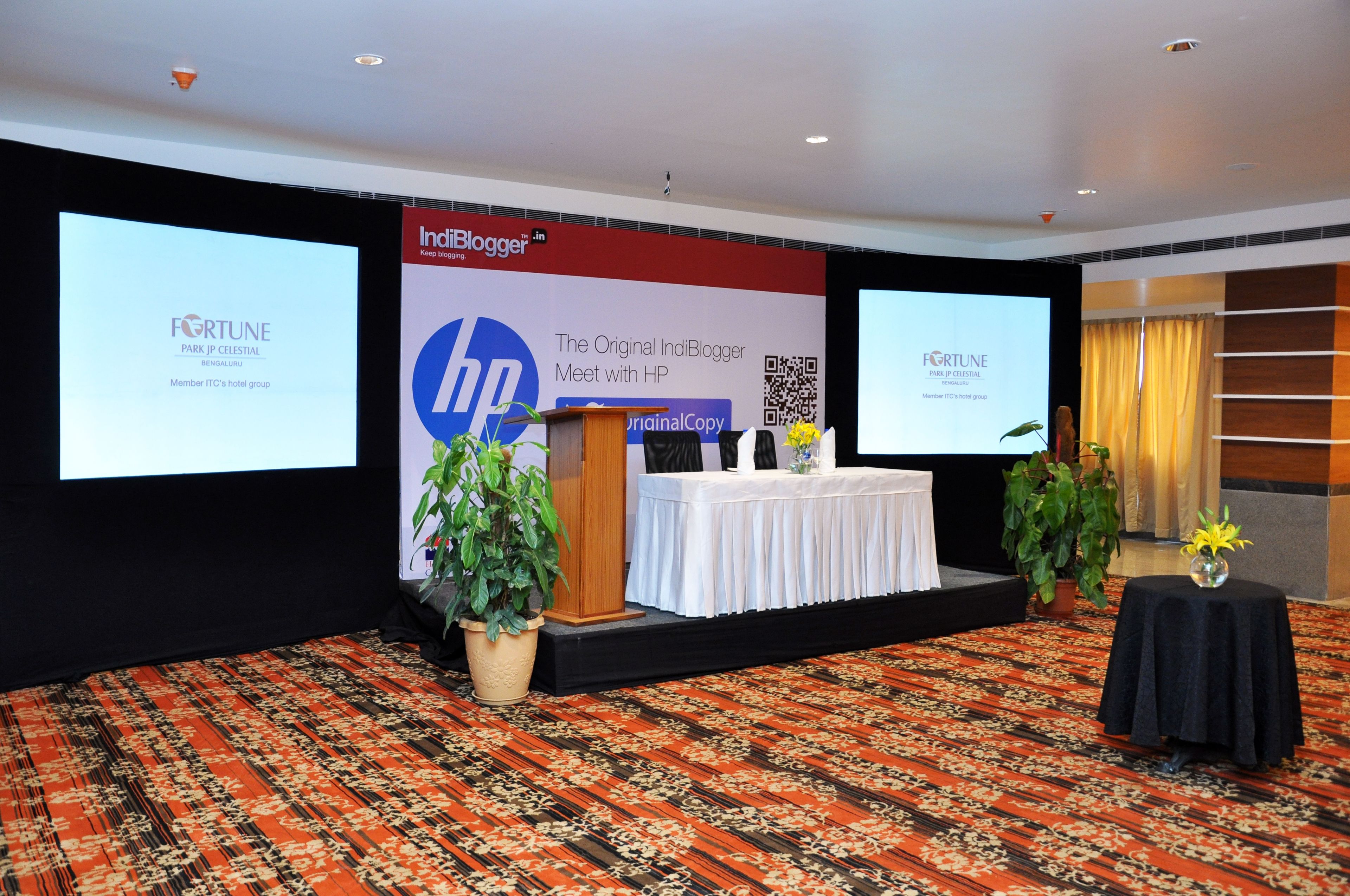 A conference room with a stage, two large screens, a podium, and potted plants.