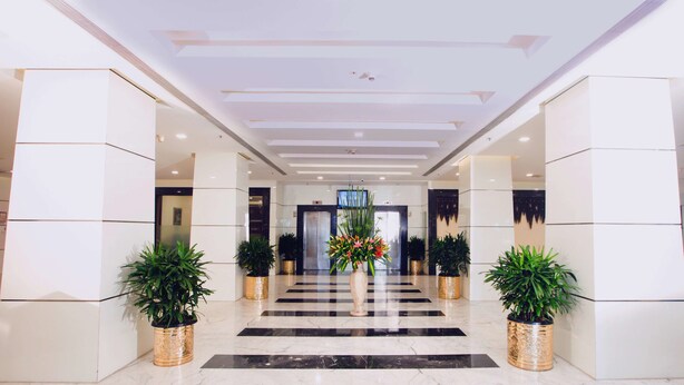 Un hall d’hôtel moderne avec un sol en marbre, deux plantes en pot, une composition florale centrale et un écran de télévision fixé au mur.