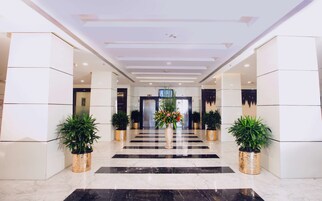 A modern hotel lobby with a marble floor, two potted plants, a central floral arrangement, and a television screen on the wall.