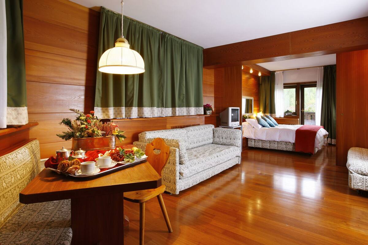 A hotel room with a wooden floor, a sofa, a dining table with a tray of food, and a television.