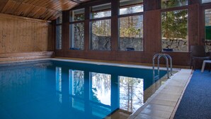 An indoor swimming pool with stone walls and wooden ceiling, large windows, and a blue floor. - Hotel Mirage (Cortina d'Ampezzo)