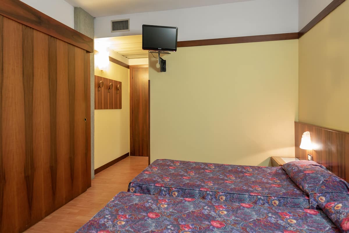 Ein Hotelzimmer mit Holzschrank, einem Bett mit floralem Bettbezug, einem Wandfernseher und einer Nachttischlampe.