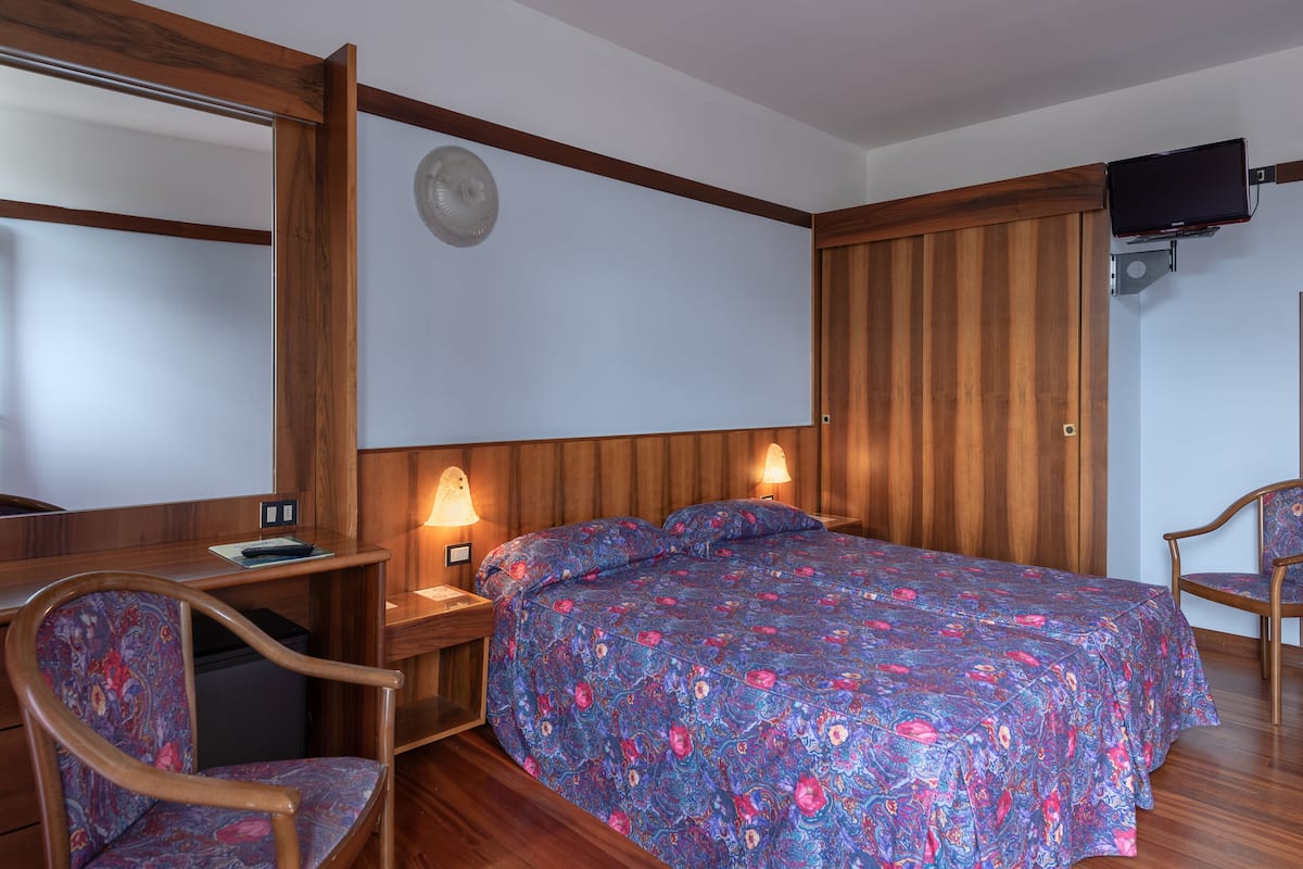 Ein Hotelzimmer mit einem hölzernen Bett, einem Schreibtisch mit Stuhl, einem an der Wand befestigten Fernseher und einem Deckenventilator.