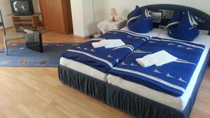 Una cama bien tendida con ropa de cama azul y blanca, una mesita de vidrio, un televisor y una lámpara en una habitación sencilla.