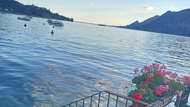 A wooden dock with metal railings overlooking a calm lake with boats and mountains in the background. - Hotel Malcesine (Malcesine)