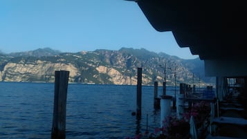 Vista sull'acqua con montagne sullo sfondo, pali di legno nell'acqua e un edificio con posti a sedere all'aperto.