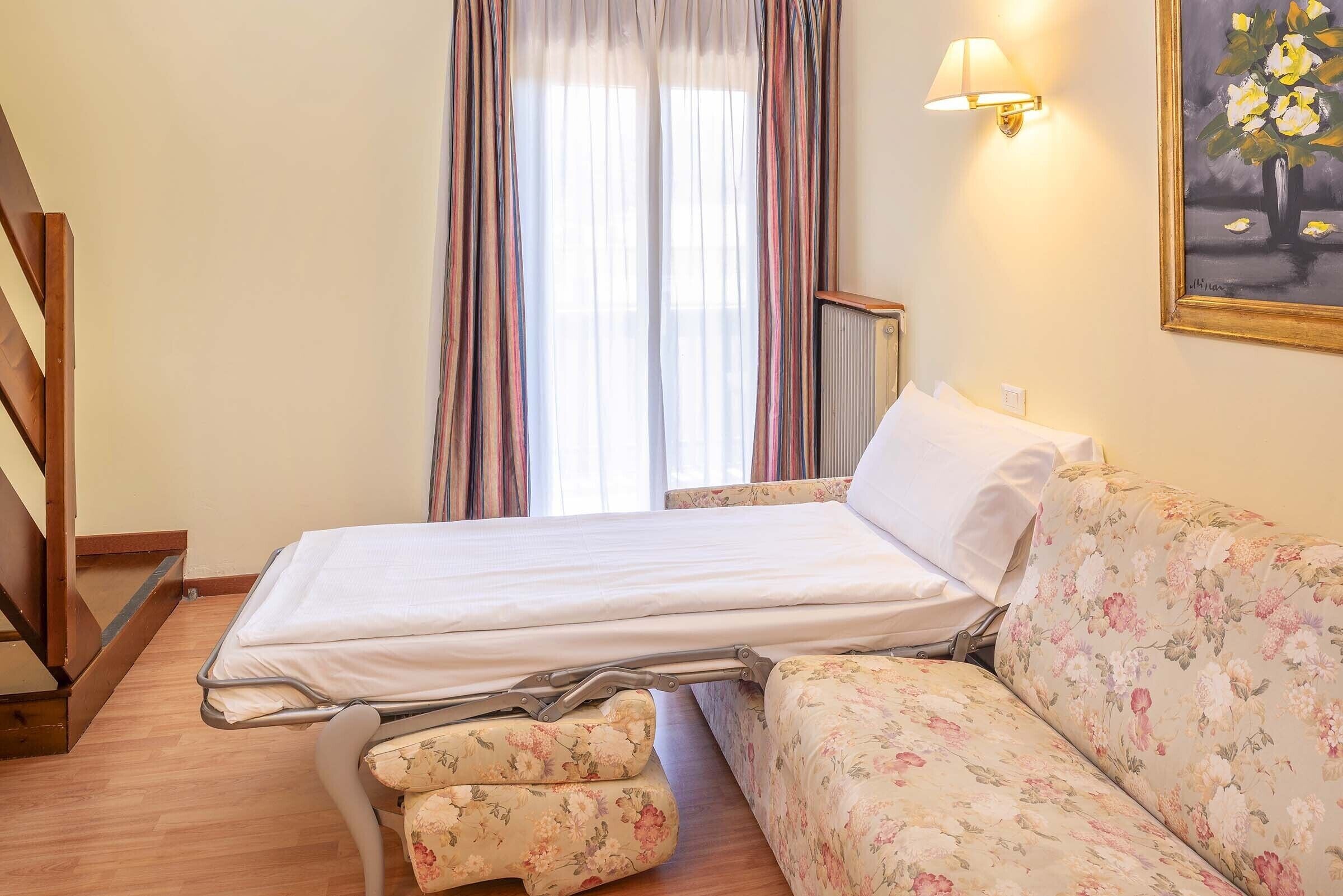 A hotel room with a bed, a sofa, a window with curtains, and a painting on the wall.
