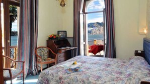 A bedroom with a bed, a desk, and a chair. There is a window with a view of the sea and mountains. - Hotel Malcesine (Malcesine)