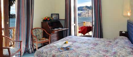 Un dormitorio con cama, escritorio y silla. Se ve una ventana con vistas al mar y a las montañas.