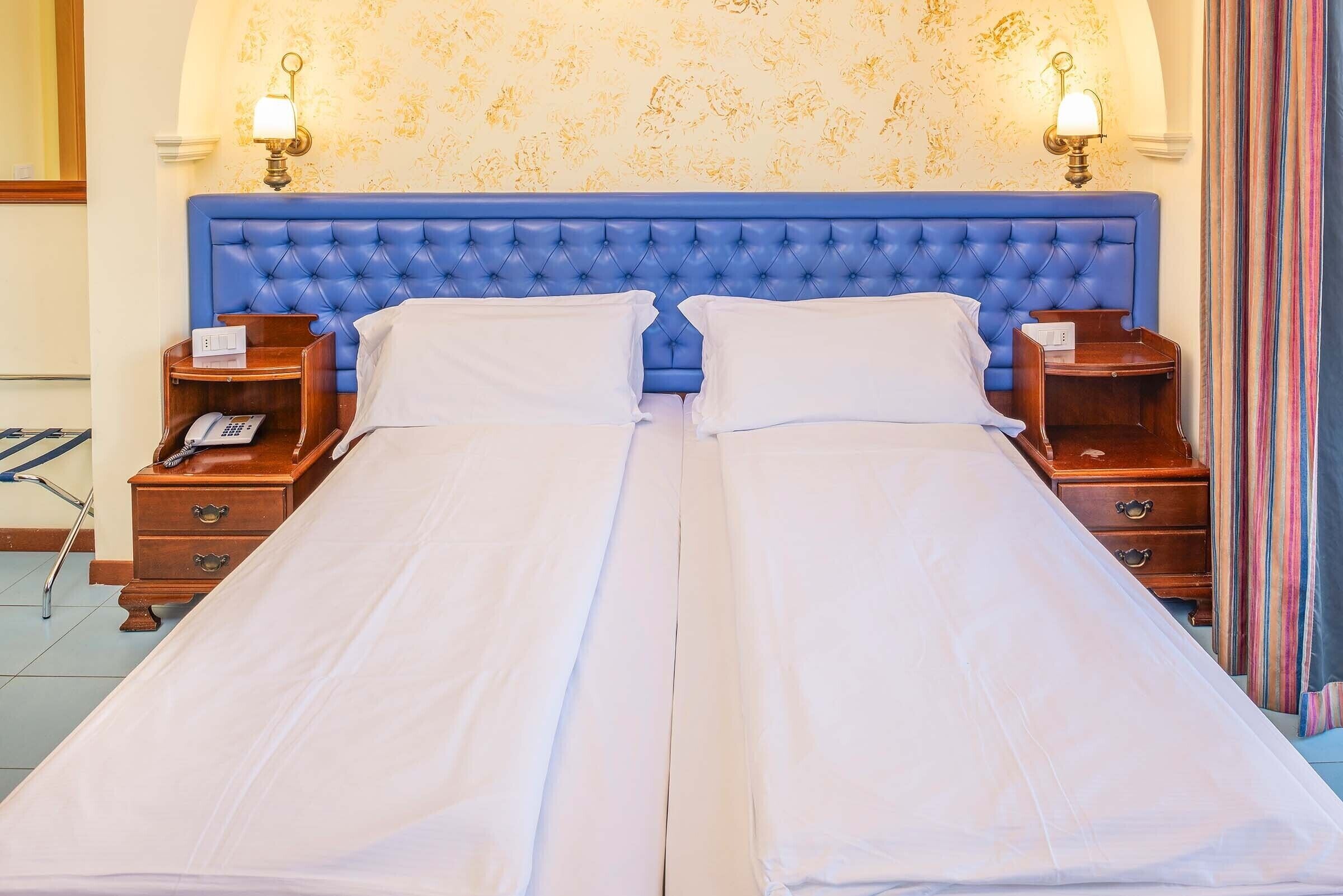 Ein Doppelbett mit einem gepolsterten Kopfteil, zwei Nachttische mit Lampen und ein Telefon auf der linken Seite.