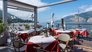 An outdoor dining area with tables and chairs, overlooking a body of water and mountains. - Hotel Malcesine (Malcesine)