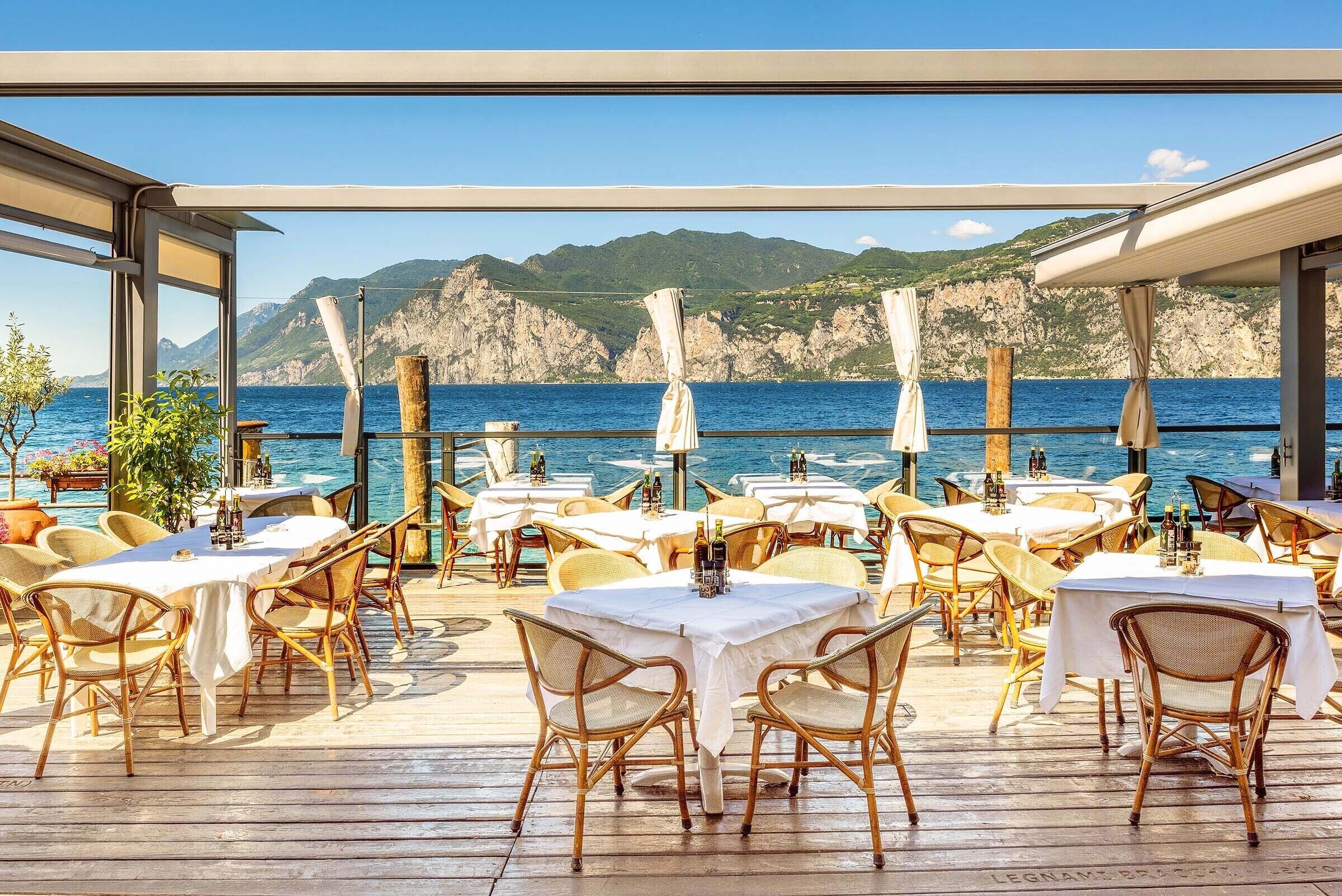 An outdoor dining area with tables and wicker chairs, overlooking a body of water and rocky cliffs.