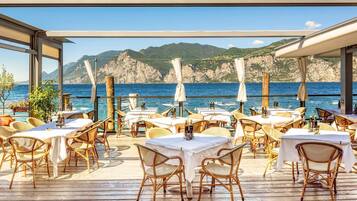 Area pranzo all'aperto con tavoli e sedie in vimini, affacciata su uno specchio d'acqua e scogliere rocciose.