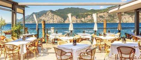 An outdoor dining area with tables and wicker chairs, overlooking a body of water and rocky cliffs.